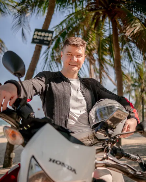 Anton Shubin smiling on his Honda motorcycle on a beach, helmet under his arm